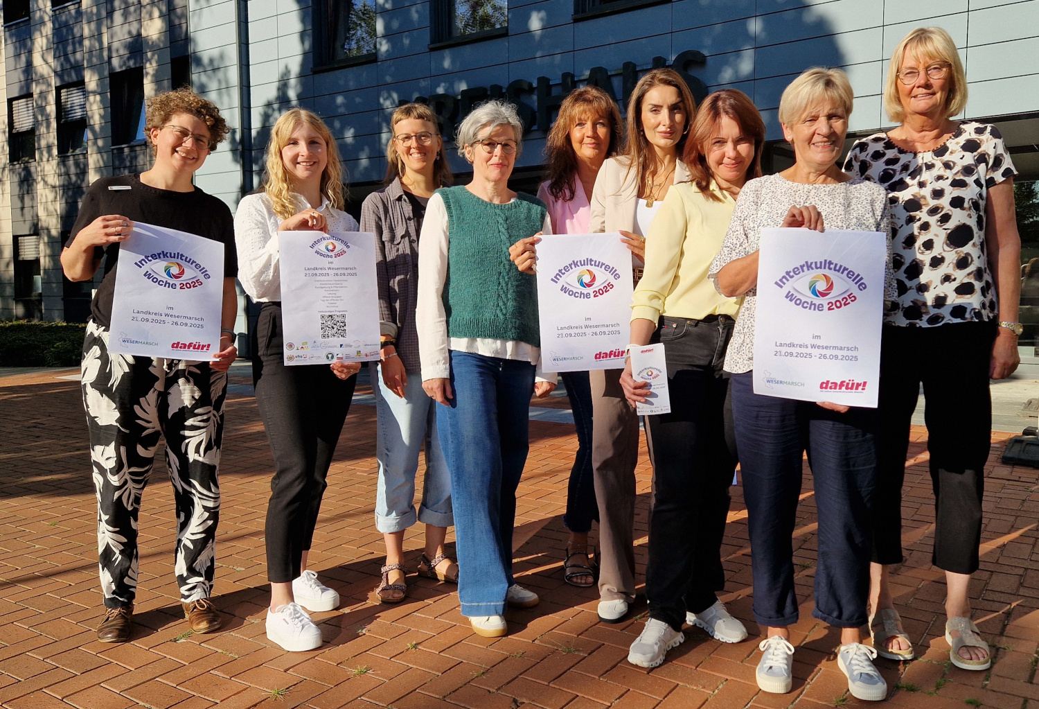 Eine Gruppe von neun Frauen steht vor dem Kreishaus Wesermarsch und hält Plakate und Flyer zur Interkulturellen Woche 2025 in die Kamera. Auf den Plakaten ist das Logo der Interkulturellen Woche sowie der Hinweis auf den Zeitraum vom 21. bis 26. September 2025 zu sehen.
