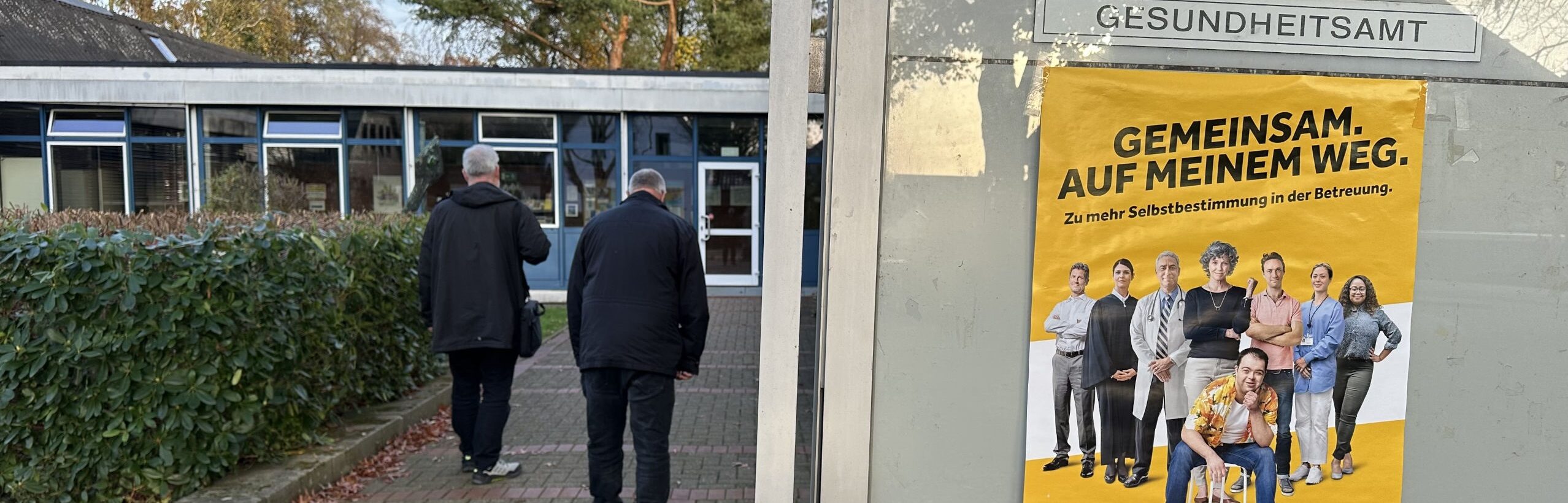 Zwei ältere Menschen auf dem Weg zur Betreuungsstelle. Daneben ein auffällig gelbes Werbeplakat zum Thema.