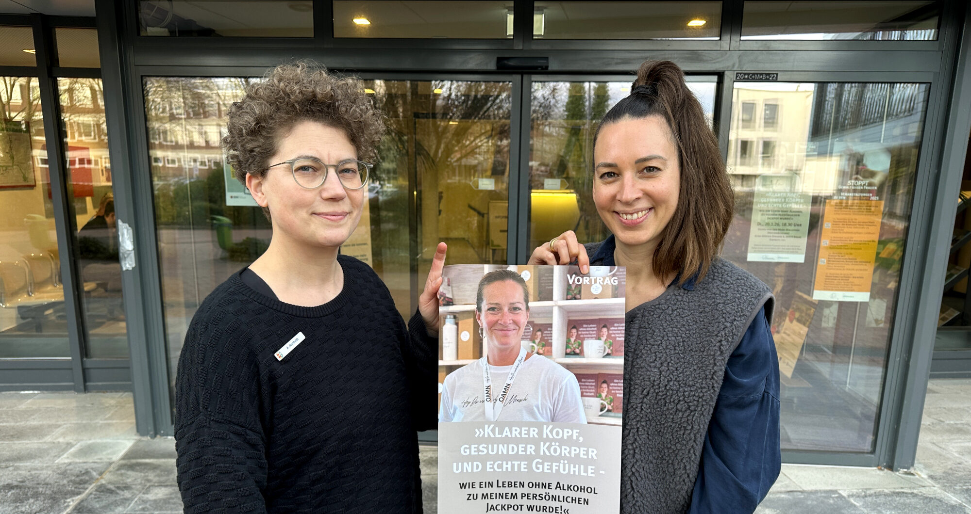 Anne Humboldt und Maren Ozanna stehen vor dem Kreishaus in Brake und präsentieren das Plakat zur Veranstaltung