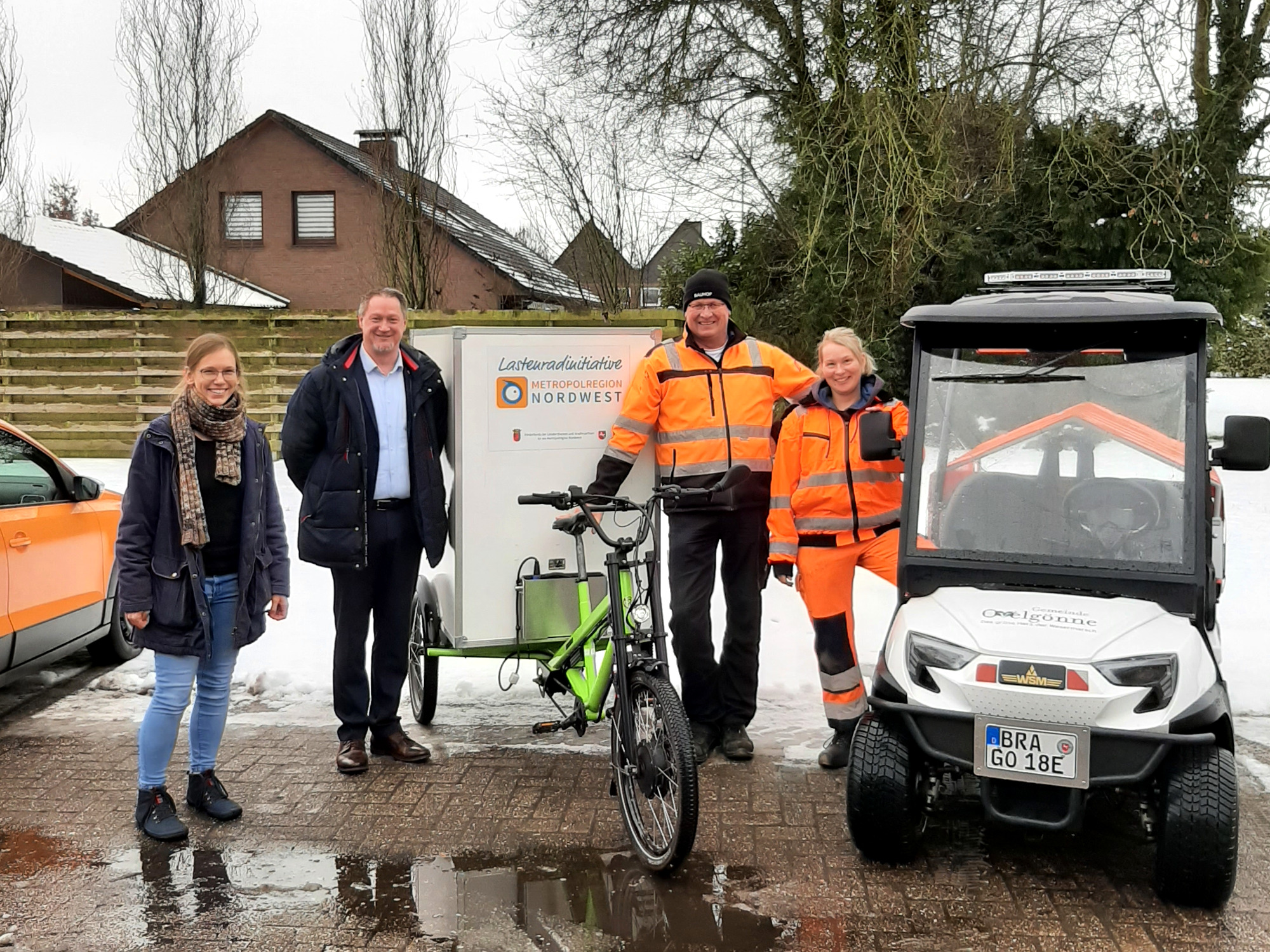 Vier Personen stehen im Freien auf einem nassen Parkplatz neben einem grünen E-Lastenrad mit Anhänger und einem kleinen weißen Elektrofahrzeug. Zwei Mitarbeitende tragen orangefarbene Warnschutzkleidung, zwei weitere Personen sind in Alltags- bzw. Businesskleidung gekleidet. Auf dem Anhänger ist ein Schild mit der Aufschrift „Lastenradinitiative Metropolregion Nordwest“ zu sehen. Im Hintergrund stehen Wohnhäuser und winterliche Bäume.