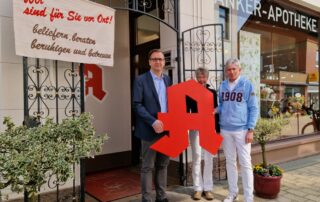 Landrat Stephan Siefken steht mit mit dem Inhaber-Apotheken-Ehepaar der Anker-Apotheke in Elsfleth und einem roten Apotheker-A in der Hand vor der Apotheke.