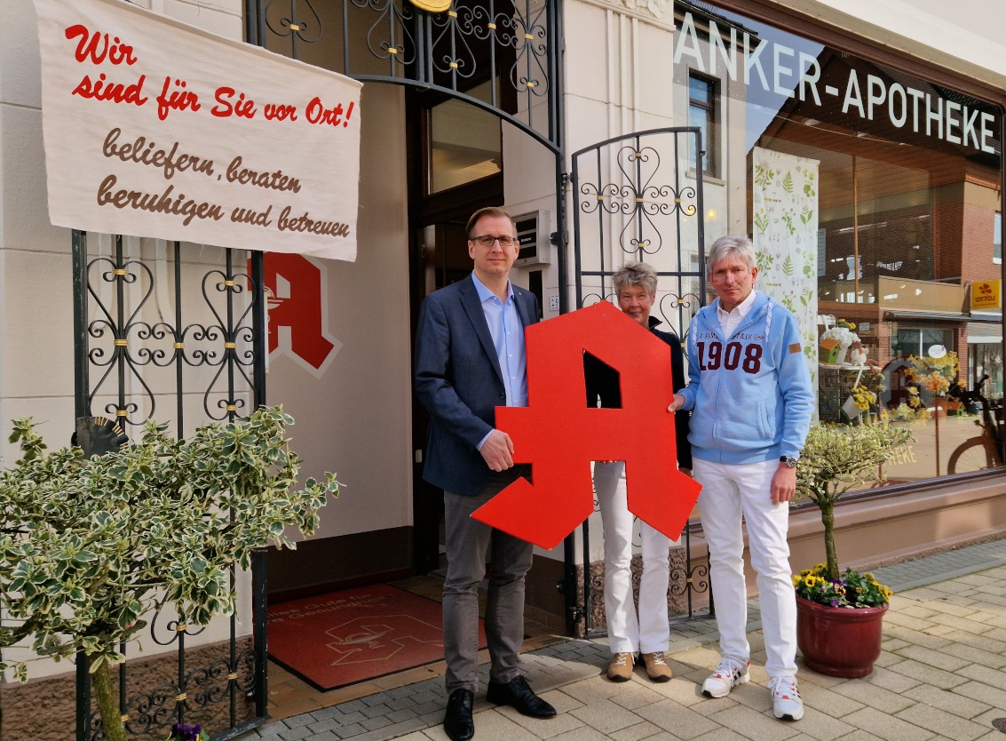 Landrat Stephan Siefken steht mit mit dem Inhaber-Apotheken-Ehepaar der Anker-Apotheke in Elsfleth und einem roten Apotheker-A in der Hand vor der Apotheke.
